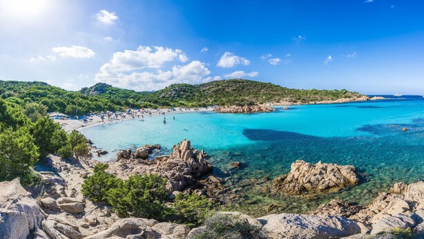 Paesaggio costiero con acque turchesi cristalline, terreno roccioso e una spiaggia.