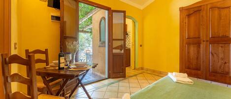 A room with a bed, a wooden chair, a table with a bottle and glasses, and a door leading outside.