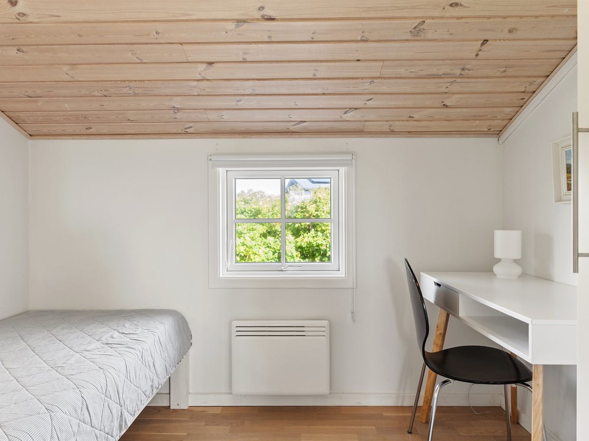 A small, bright room with a wooden ceiling, a bed, a desk with a chair, and a window with a view of greenery.