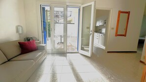 Ein Wohnzimmer mit einer Couch, einer Tür, die auf einen Balkon führt, und einer durch die Tür sichtbaren Küche.
