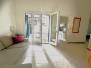 Ein Wohnzimmer mit einer Couch, einer Tür, die auf einen Balkon führt, und einer durch die Tür sichtbaren Küche.