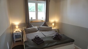 Un letto ben rifatto con cuscini e asciugamani piegati, un comodino con una lampada e una finestra con tende.