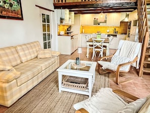 Ein gemütliches Wohnzimmer mit einer Couch, einem Couchtisch und einem Sessel. Im Hintergrund befindet sich eine Küche mit einem Esstisch und Stühlen.