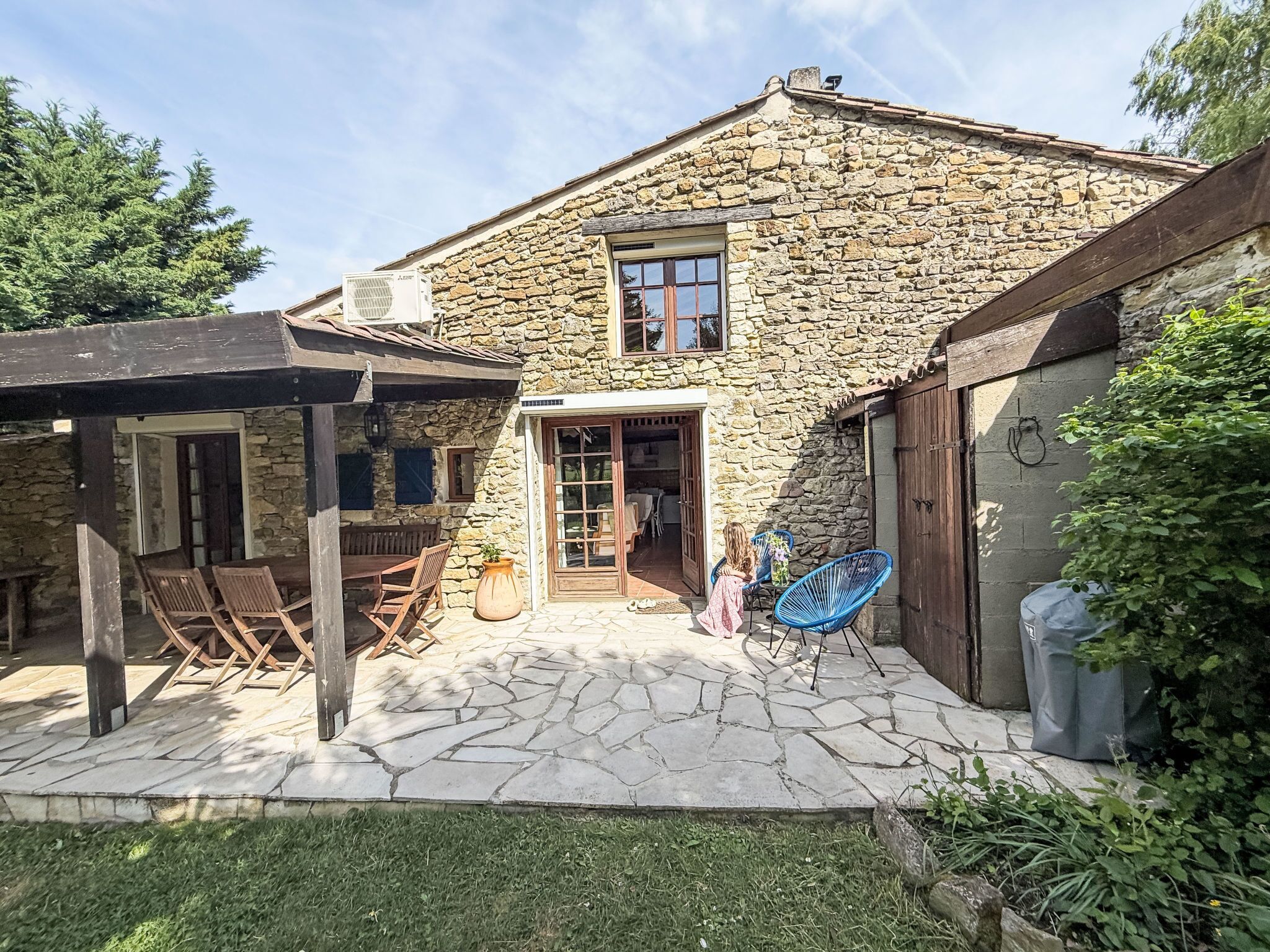 A stone house with a covered patio, a wooden table and chairs, and a child playing.