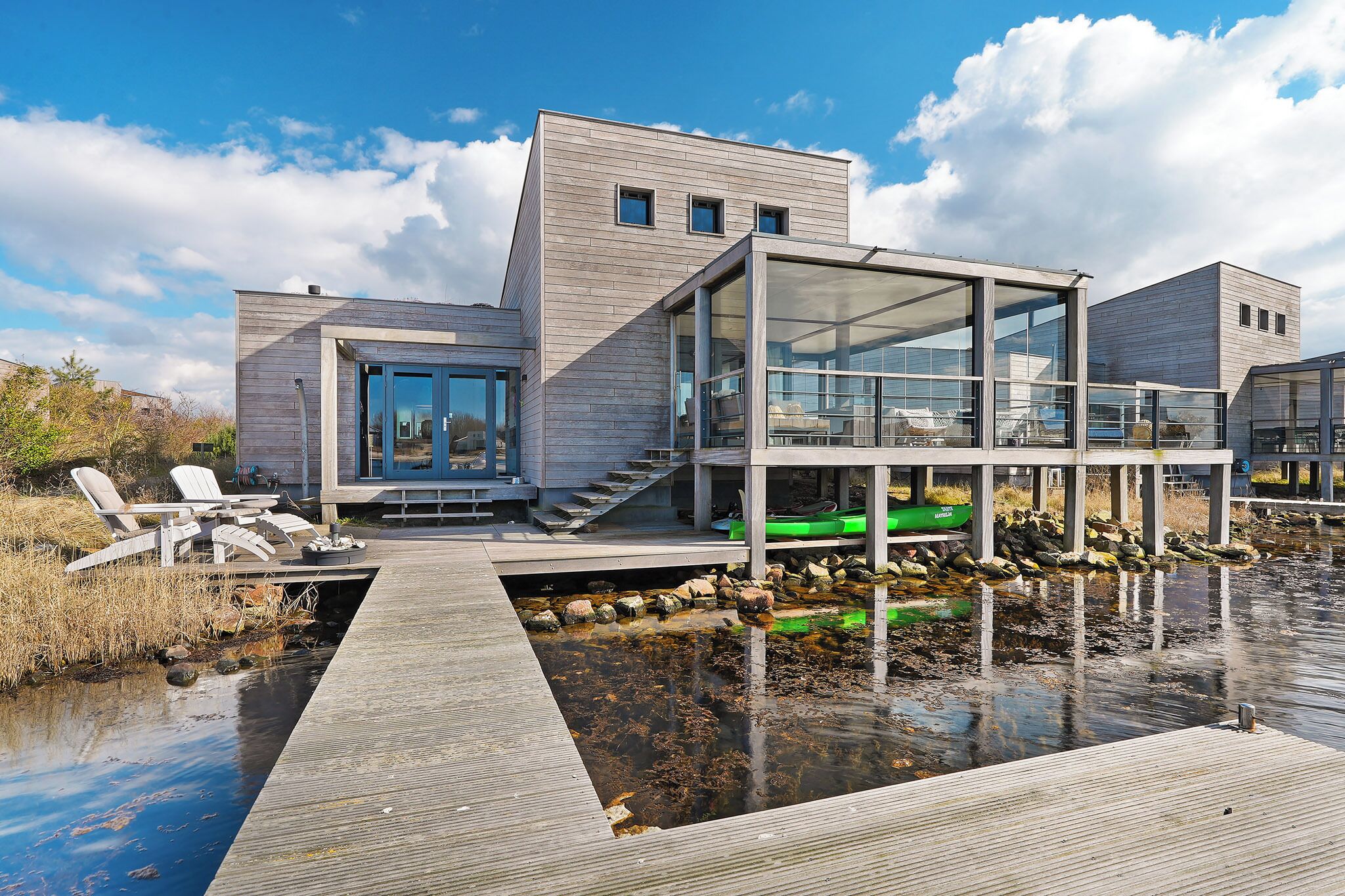 Une maison moderne avec une façade en bois, une véranda en verre et une jetée qui s’avance sur une étendue d’eau.