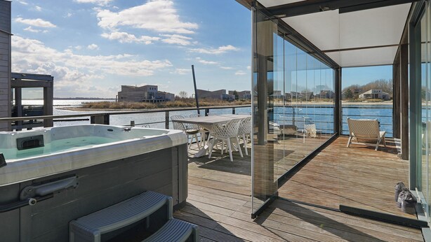 A deck with a hot tub, outdoor seating, and a glass wall offering a view of the water and buildings. - The terraces of a 4-person Villa Super VIP are located directly by the water, with access to a private swimming pier, jacuzzi and Infrared sauna. (Ouddorp)
