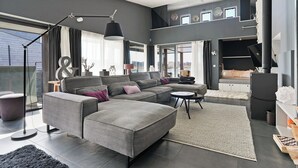 A modern living room with a grey sectional sofa, a round coffee table, and a black floor lamp. - The terraces of a 4-person Villa Super VIP are located directly by the water, with access to a private swimming pier, jacuzzi and Infrared sauna. (Ouddorp)