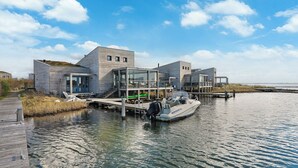 A modern waterfront property with a dock, a boat, and a glass-enclosed structure. - The terraces of a 4-person Villa Super VIP are located directly by the water, with access to a private swimming pier, jacuzzi and Infrared sauna. (Ouddorp)
