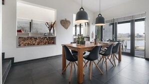A modern dining area with a wooden table and black chairs, large windows with a view of the water, and a built-in shelf with firewood. - The terraces of a 4-person Villa Super VIP are located directly by the water, with access to a private swimming pier, jacuzzi and Infrared sauna. (Ouddorp)