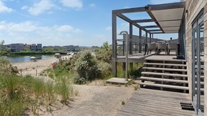 A modern building with a wooden deck and outdoor seating area overlooking a beach and water. - The 4-person Villa Super VIP features a luxurious and comfortably furnished interior with a direct waterfront location, various terraces offering unobstructed views over the water. (Ouddorp)