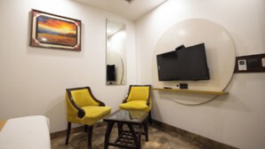 A room with two yellow chairs, a small table, a flat-screen TV mounted on a circular wall, and a painting of a sunset. - Hotel Namoh (New Delhi)