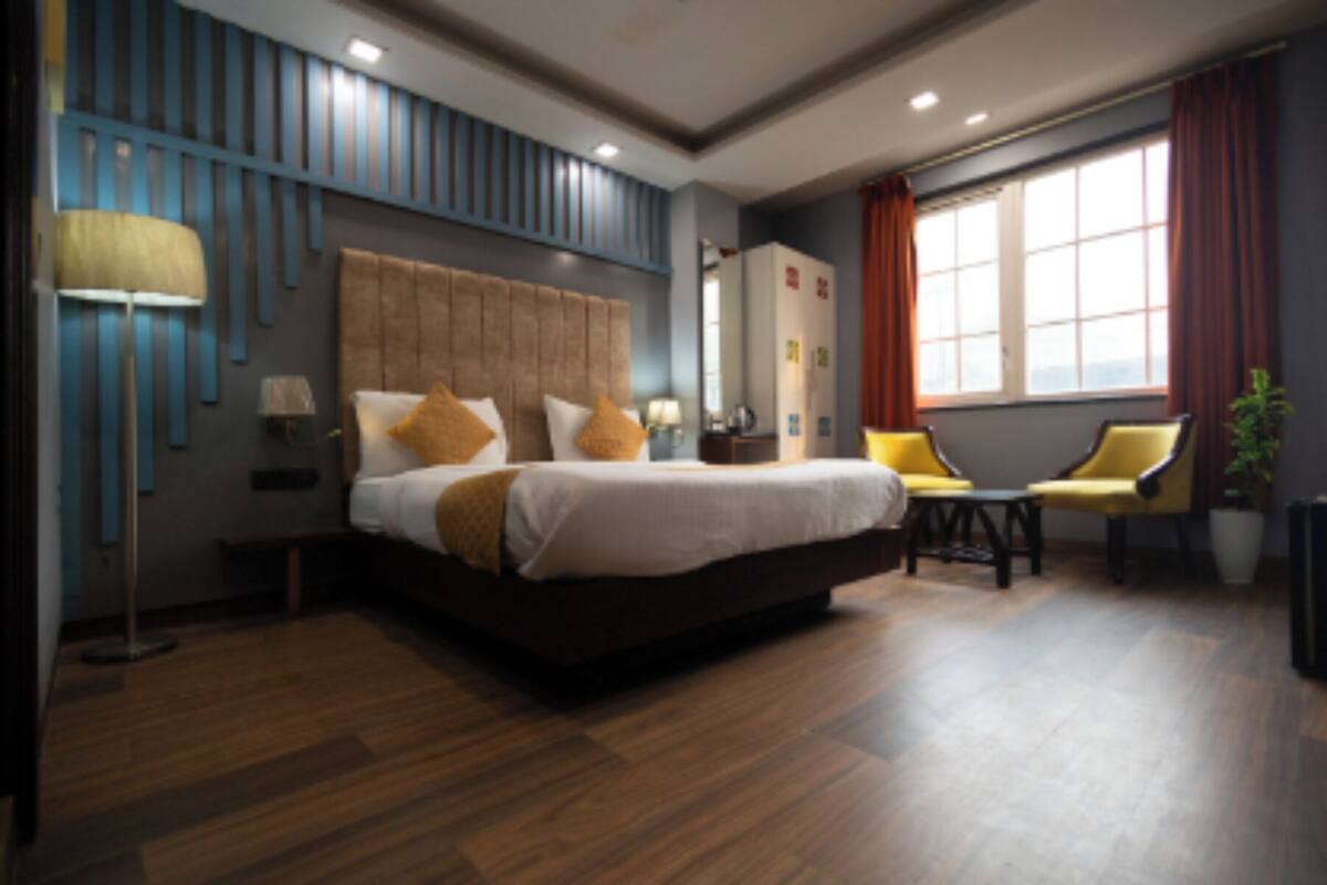 A hotel room with a large bed, a desk, two chairs, a potted plant, and a window with curtains.