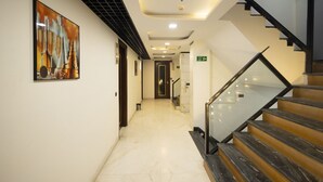 A modern hotel hallway with a glass railing staircase, a framed artwork on the wall, and a clean, tiled floor. - Hotel Namoh (New Delhi)