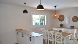 A modern kitchen with white cabinetry, a wooden countertop, and a dining area with white chairs. There are pendant lights above the kitchen and a window with a view of greenery outside.