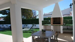 An outdoor patio with a table and chairs, a barbecue grill, and a view of a neatly trimmed hedge and lawn.