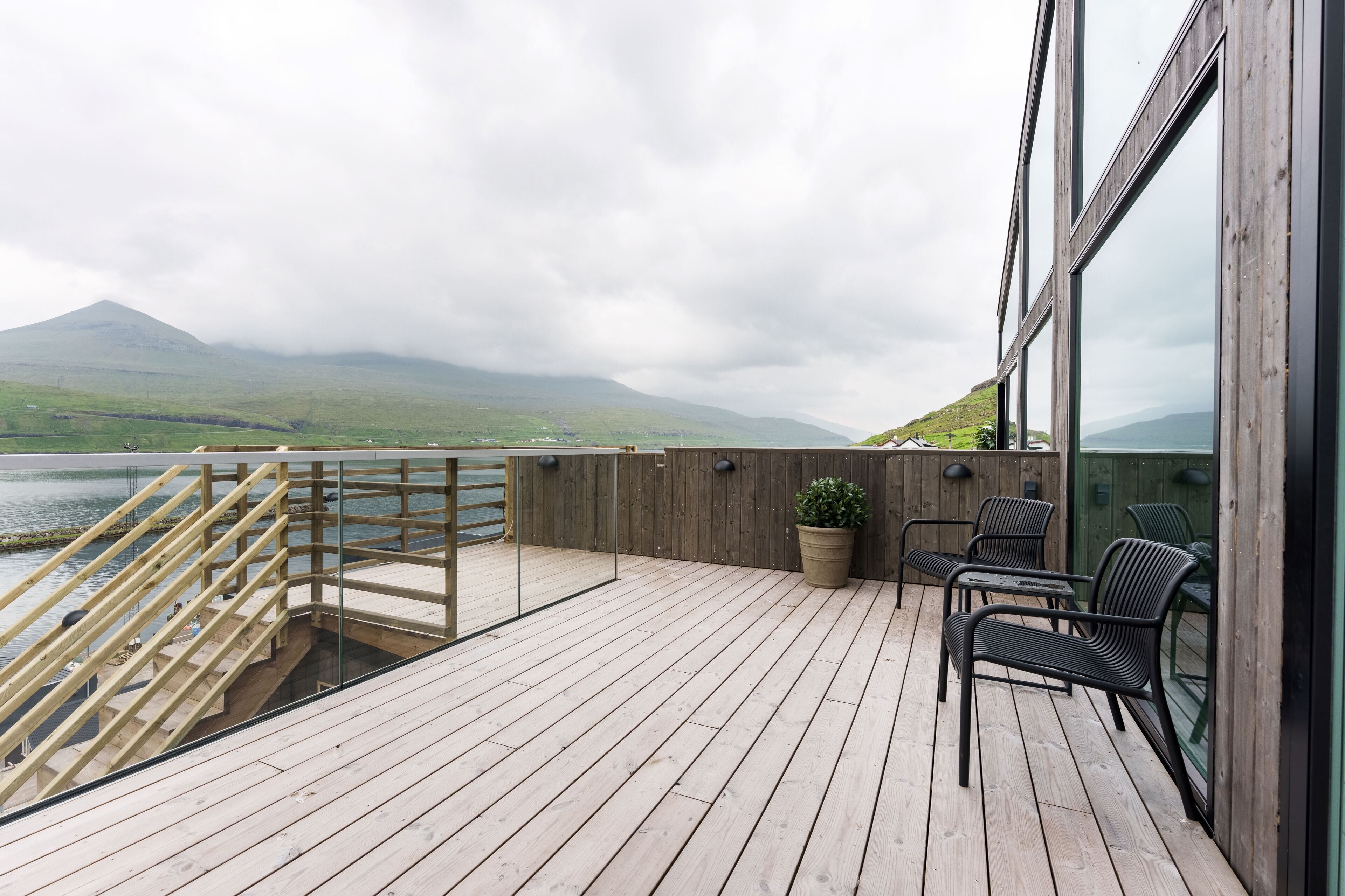A wooden deck with a glass balustrade, outdoor seating, and a view of a mountain and lake.