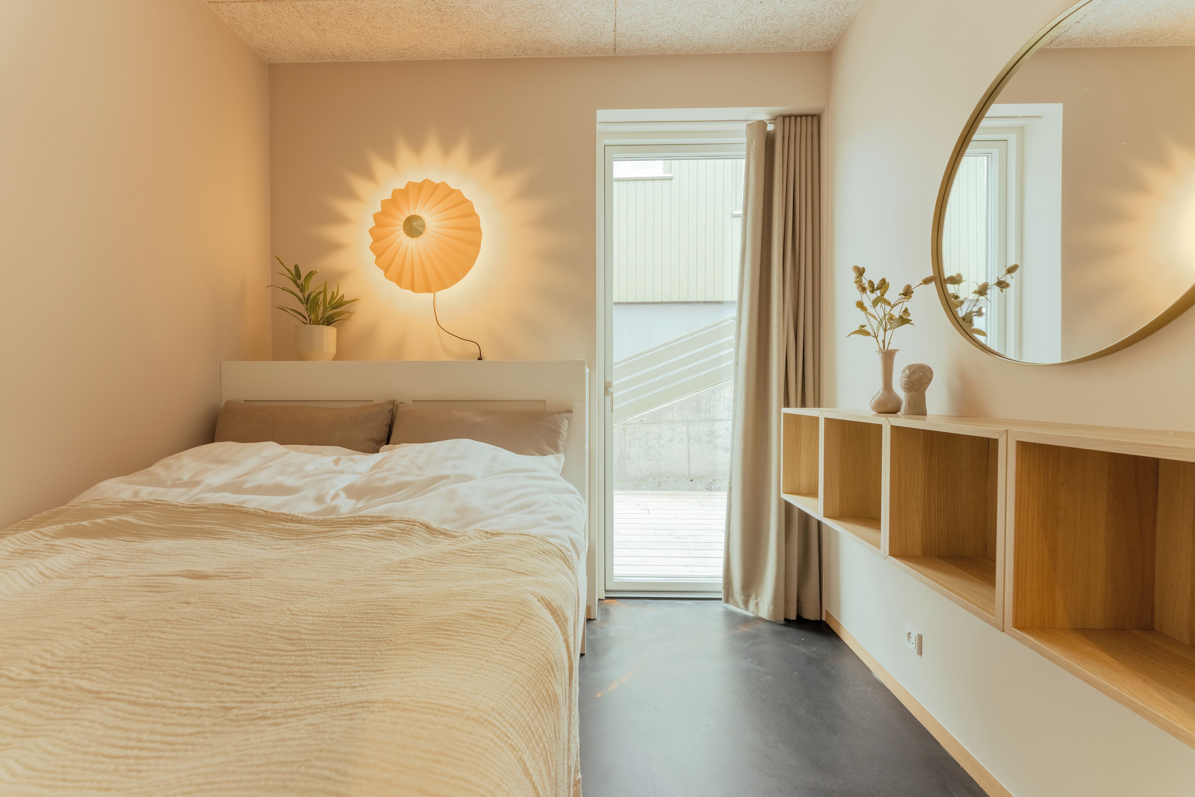 A bedroom with a bed, a wall-mounted lamp, a mirror, and a sliding door leading to a balcony.