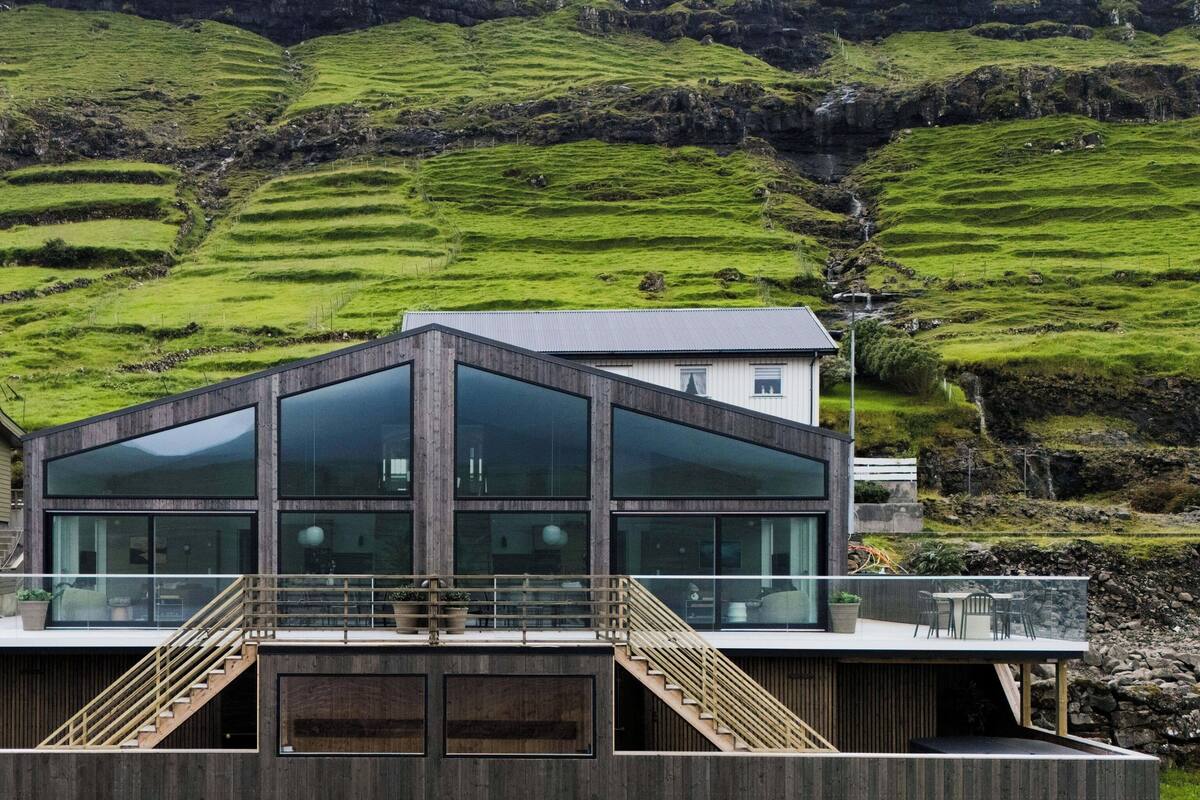 Ein modernes Gebäude mit großen Glasfenstern, Holzfassade und einer Treppe zu einer Terrasse mit Sitzgelegenheiten.