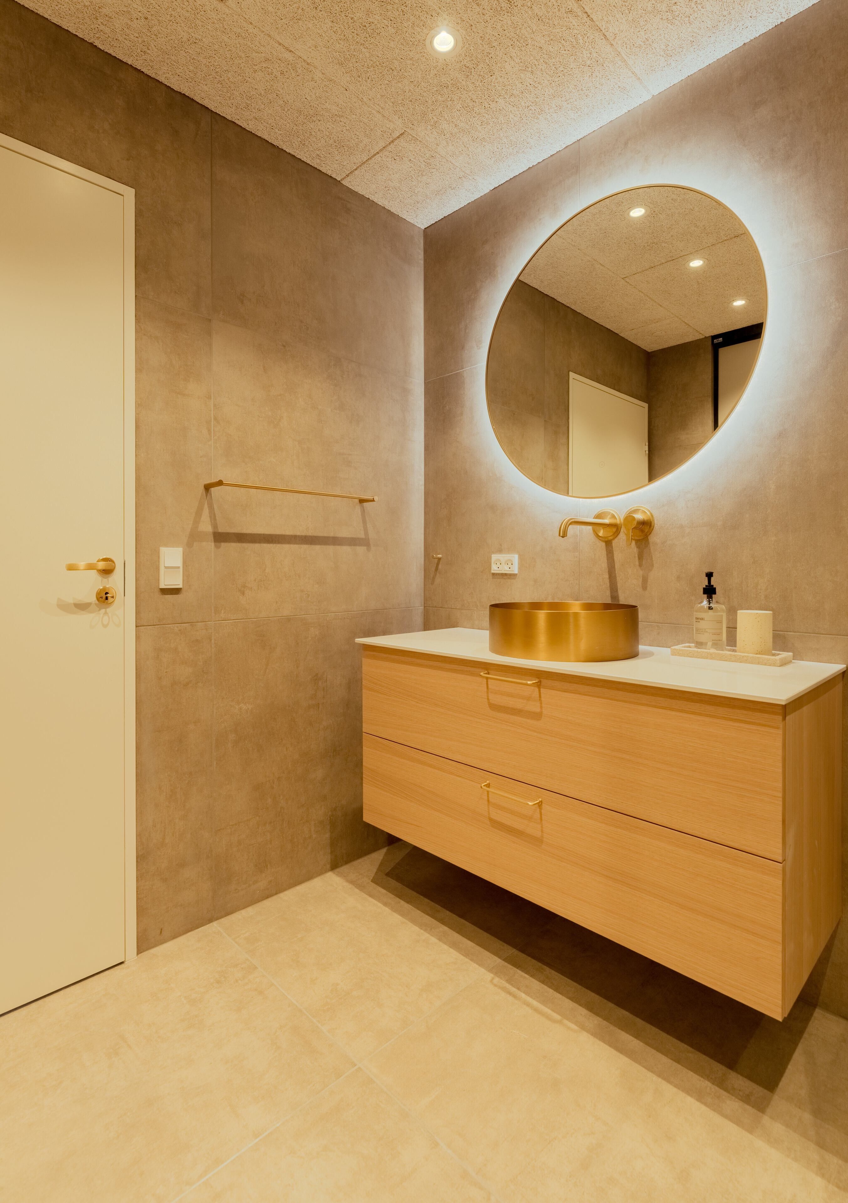 Un baño moderno con un espejo redondo, tocador de madera y lavamanos dorado.