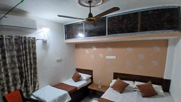 A hotel room with two single beds, a ceiling fan, an air conditioning unit, a window with curtains, and a small table with a chair. - Kaashvi Home Stay (Varanasi)