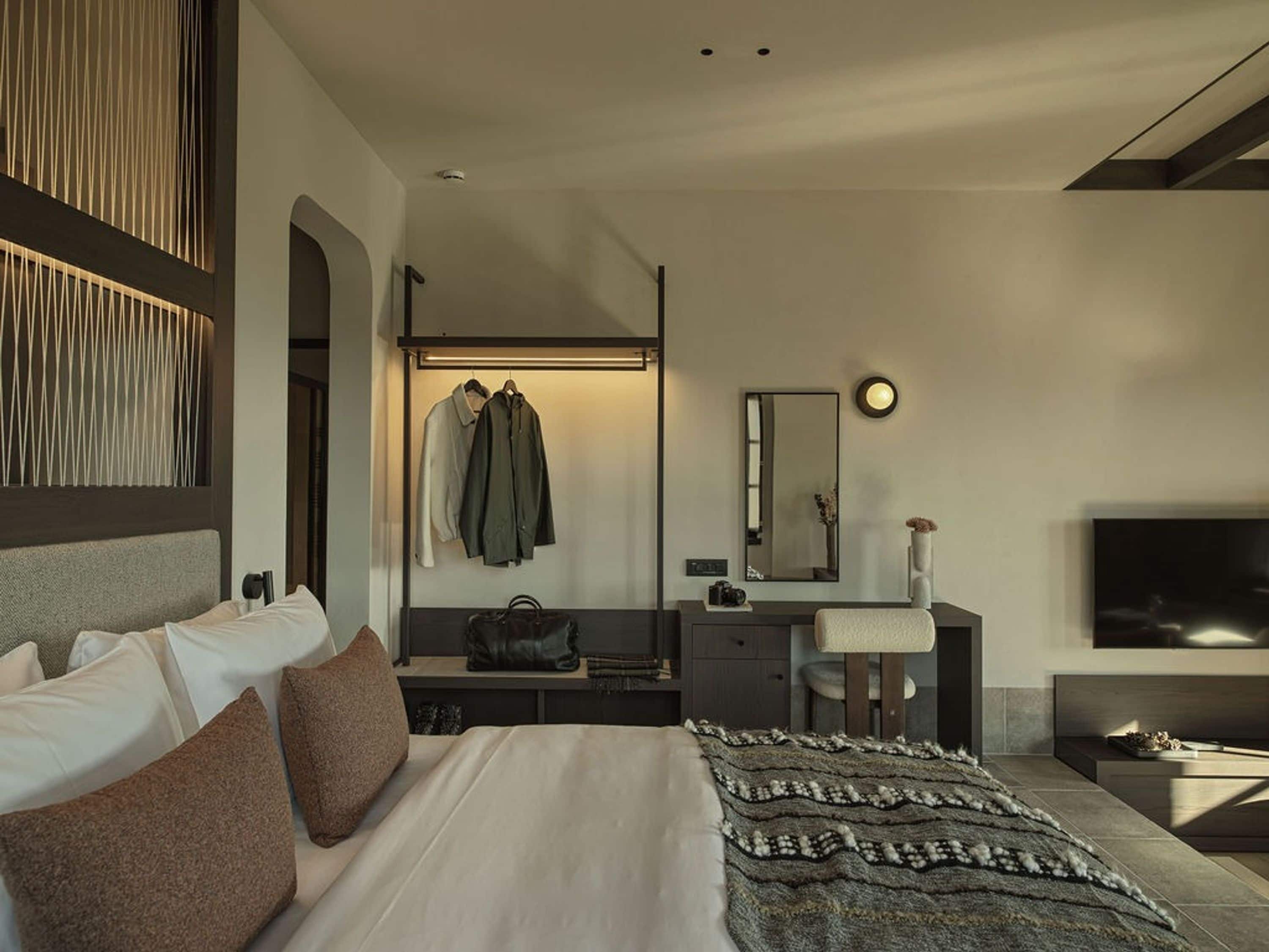 A modern hotel room with a large bed, a bedside table, a vanity with a mirror, and a flat-screen TV.