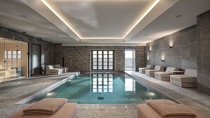 A modern indoor swimming pool with a clear view of the city through large windows, a seating area with towels, and a stone wall design. - Oreno Mountain Retreat Hotel (Zagori)