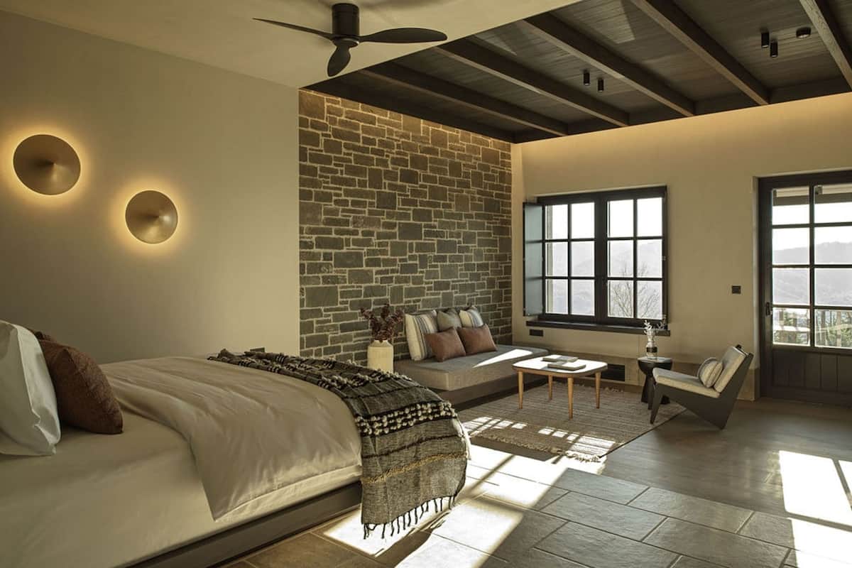 A modern bedroom with a large bed, a stone accent wall, a window with a view, and a ceiling fan.