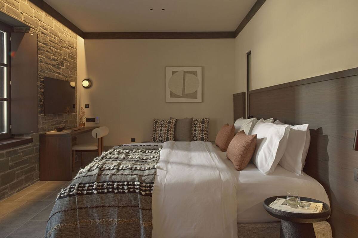 A modern bedroom with a stone accent wall, a large bed with multiple pillows, a bedside table with a magazine, and a wall-mounted clock.