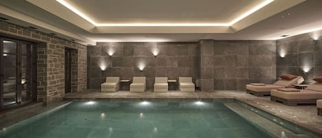 A modern indoor pool area with a clear pool, stone walls, and lounge seating.