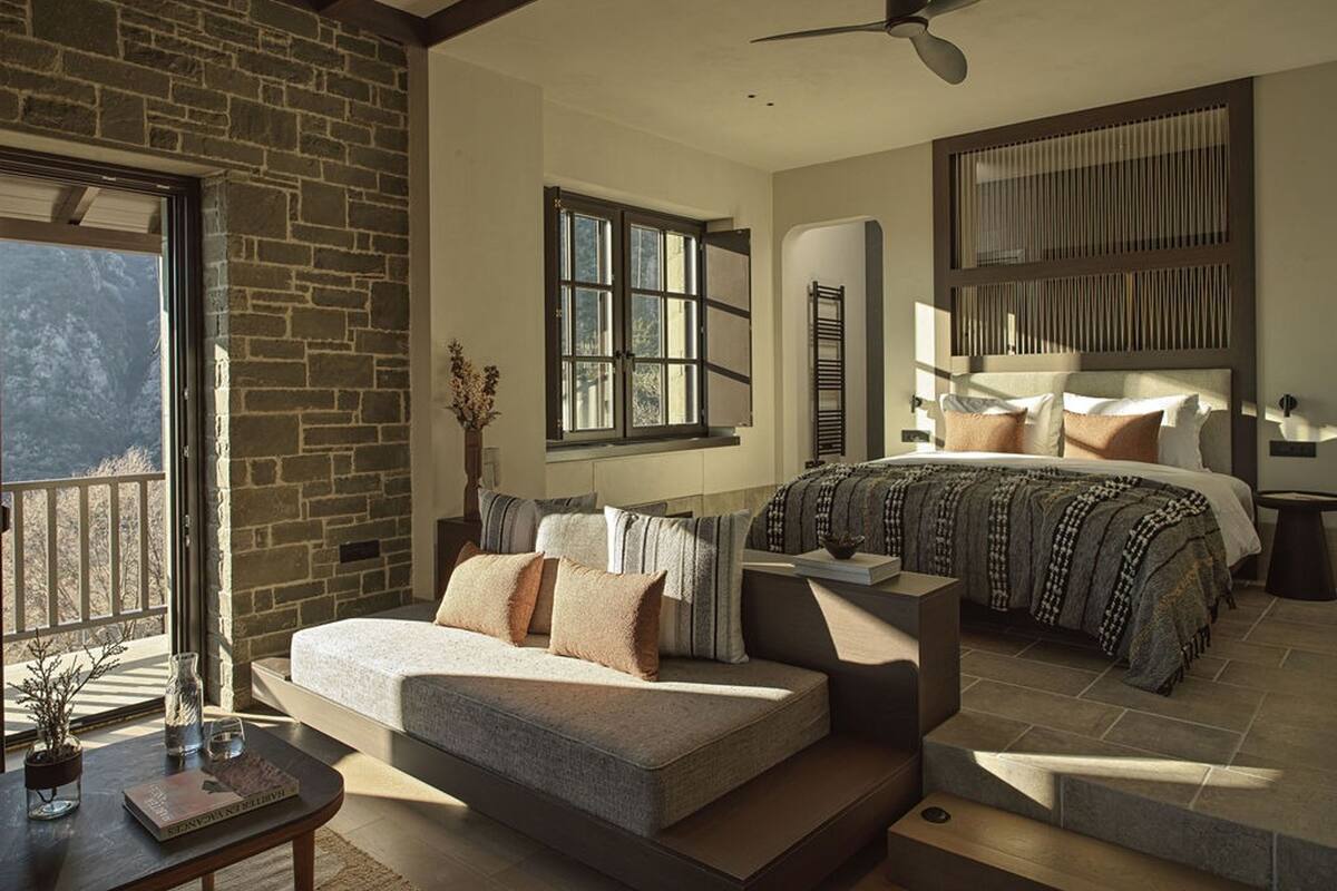A modern bedroom with a stone wall, a large bed with patterned bedding, a sofa, and a dining table with a vase and book.