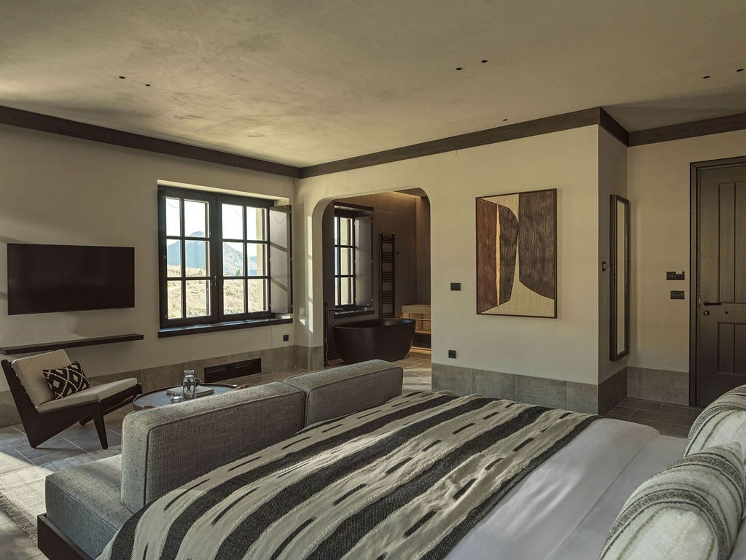 Une chambre d’hôtel moderne dotée d’un grand lit, d’un coin salon avec un canapé et des fauteuils, d’un téléviseur à écran plat fixé au mur et d’une grande fenêtre offrant une vue sur l’extérieur.