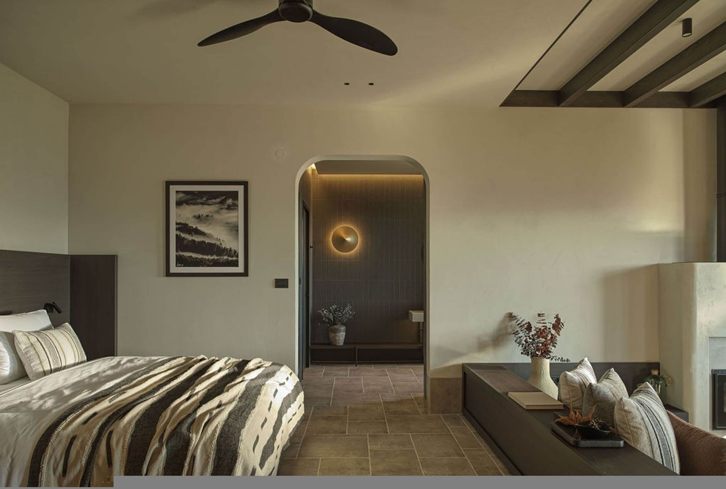 A modern bedroom with a bed, a ceiling fan, a framed picture on the wall, and a small seating area with a plant.