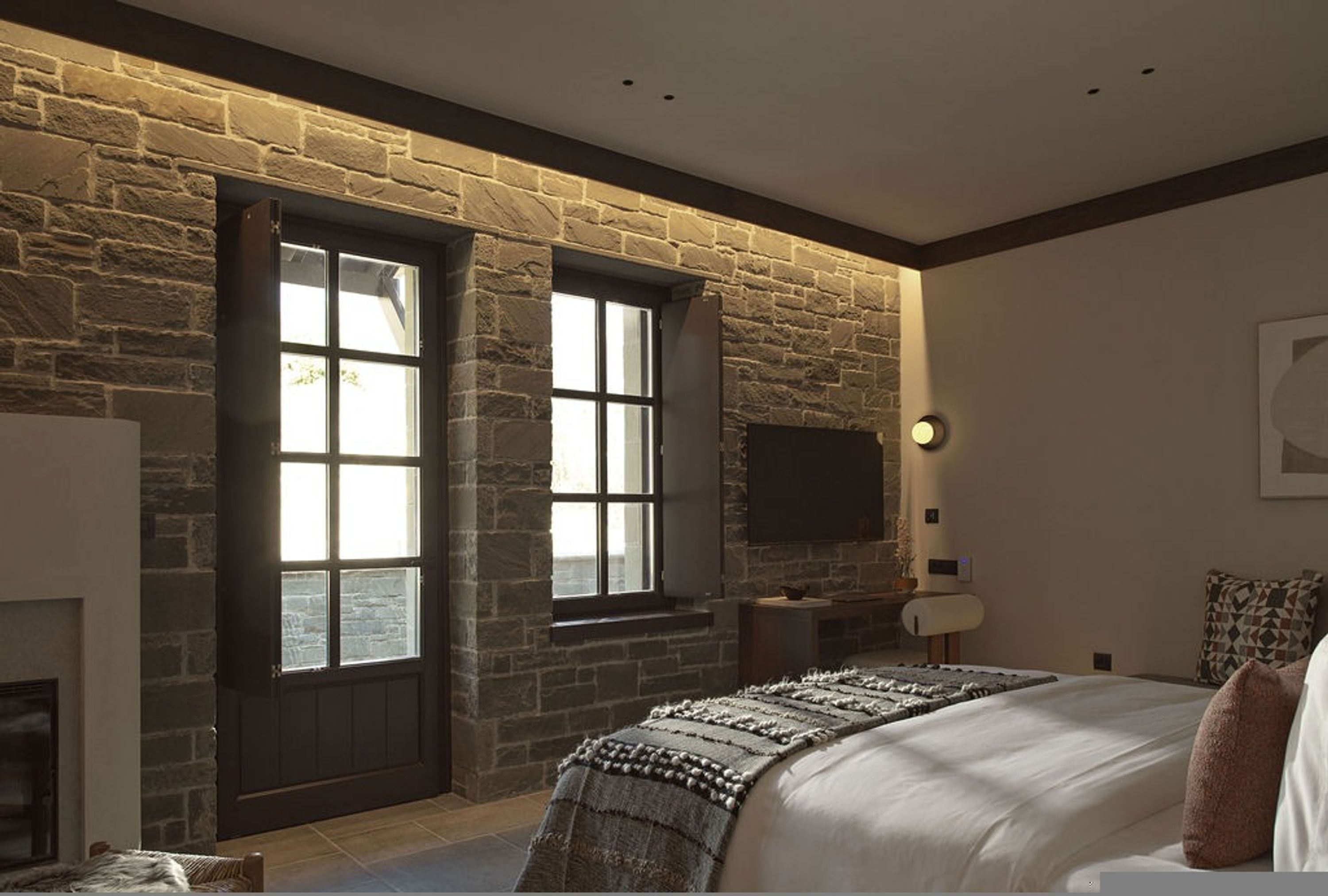 Une chambre avec un mur en pierre, une cheminée, un lit avec une couverture, une table de chevet et un téléviseur fixé au mur.