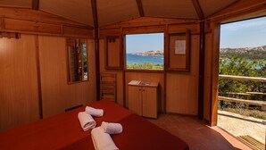 A wooden cabin room with a bed, a small cabinet, a window with a view of the sea, and a balcony with a railing.