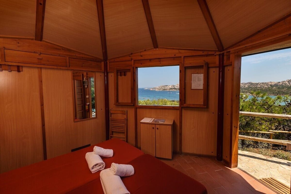 A wooden cabin room with a bed, a small cabinet, a window with a view of the sea, and a balcony with a railing.