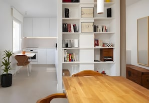 Une cuisine moderne avec des meubles blancs, une table à manger en bois et une bibliothèque contenant des livres et des objets décoratifs.