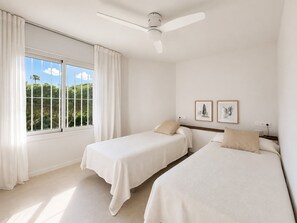 A bedroom with two beds, a ceiling fan, a window with curtains, and two framed pictures on the wall.