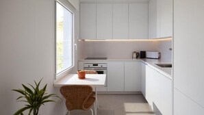 Une cuisine compacte avec des meubles blancs, une petite table à manger, une chaise, une plante en pot et une fenêtre qui laisse entrer la lumière naturelle.
