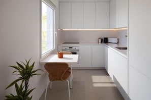 Une cuisine compacte avec des meubles blancs, une petite table à manger, une chaise, une plante en pot et une fenêtre qui laisse entrer la lumière naturelle.