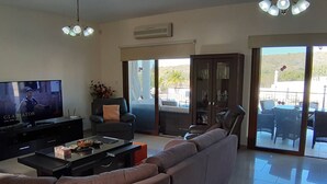 Una sala de estar con un televisor de pantalla plana grande, un sofá, una mesa de centro y una silla. Hay una puerta corrediza de vidrio que da a un patio al aire libre.