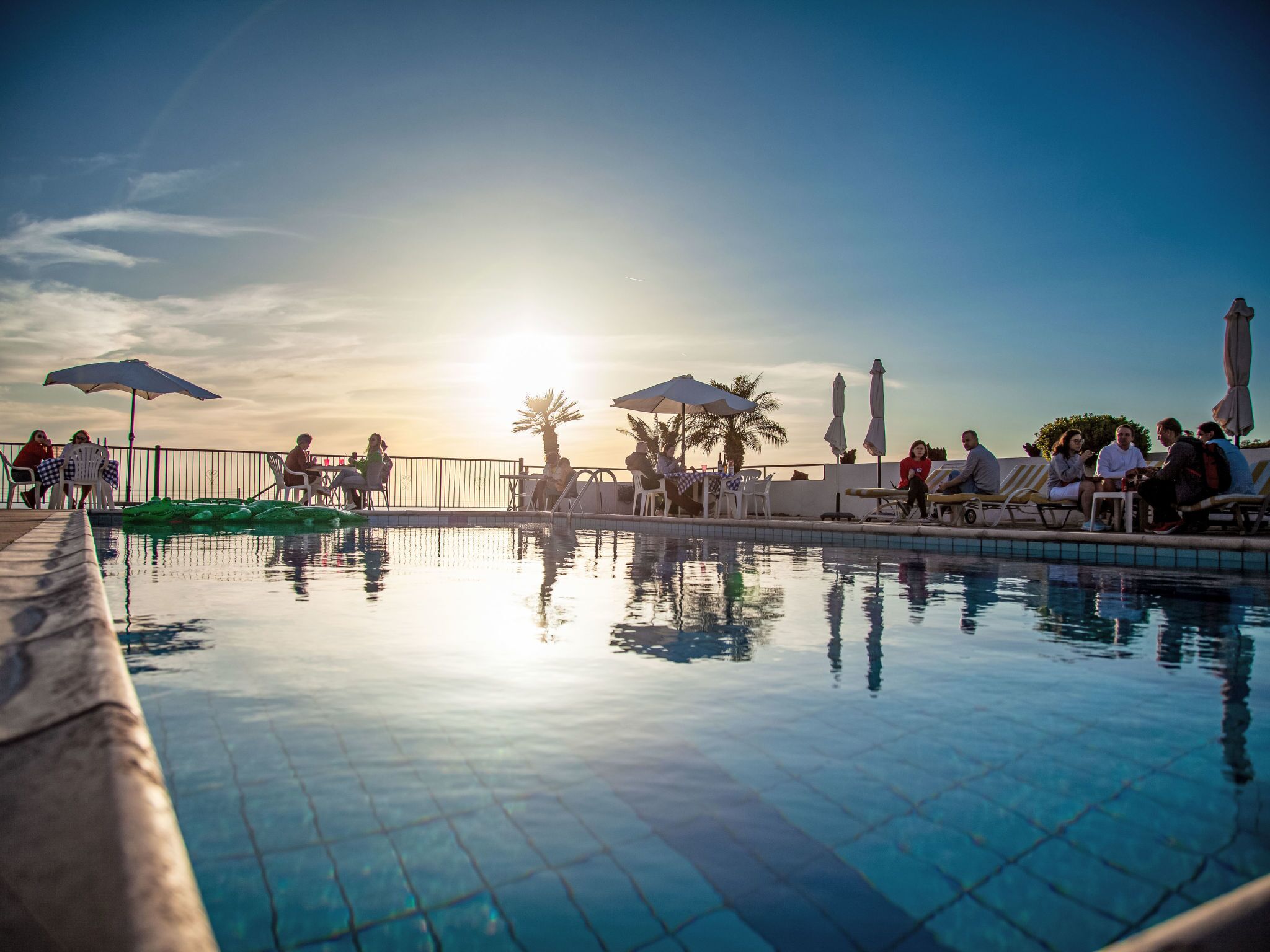 Área à beira da piscina com espreguiçadeiras e guarda-sóis, cercada por uma grade, e palmeiras ao fundo.