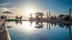 Un área junto a la piscina con sillones de descanso y sombrillas, rodeada por una valla y palmeras al fondo.