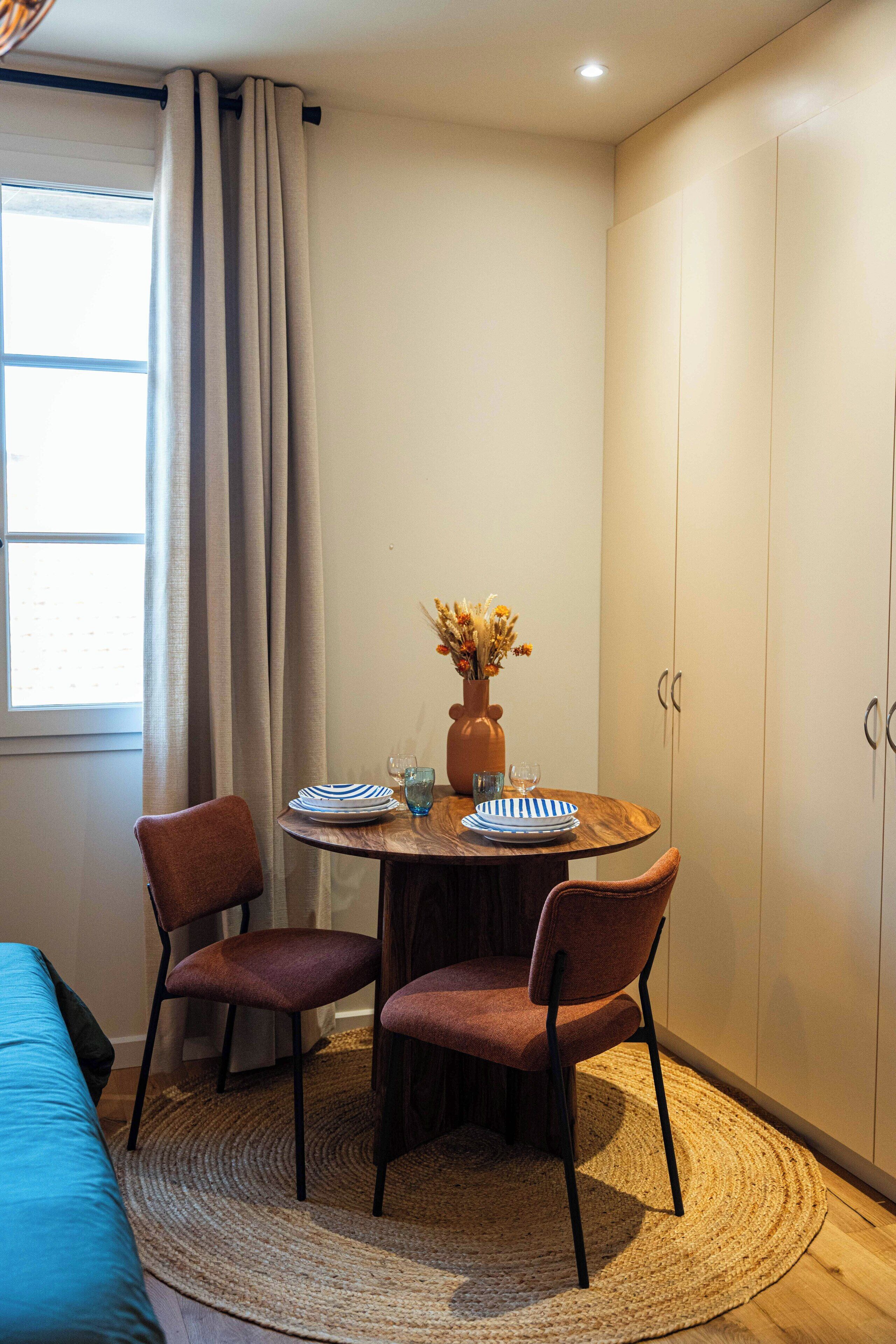A small dining area with a round table, two chairs, a vase with flowers, and a window with curtains.