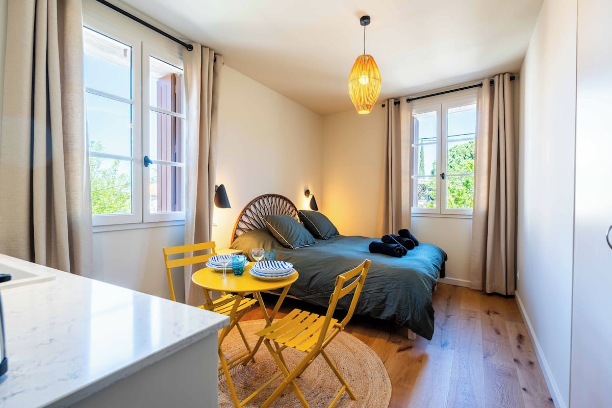 Ein gemütliches Schlafzimmer mit Bett, einem kleinen Esstisch mit Stühlen, einer Pendelleuchte und großen Fenstern mit Vorhängen.