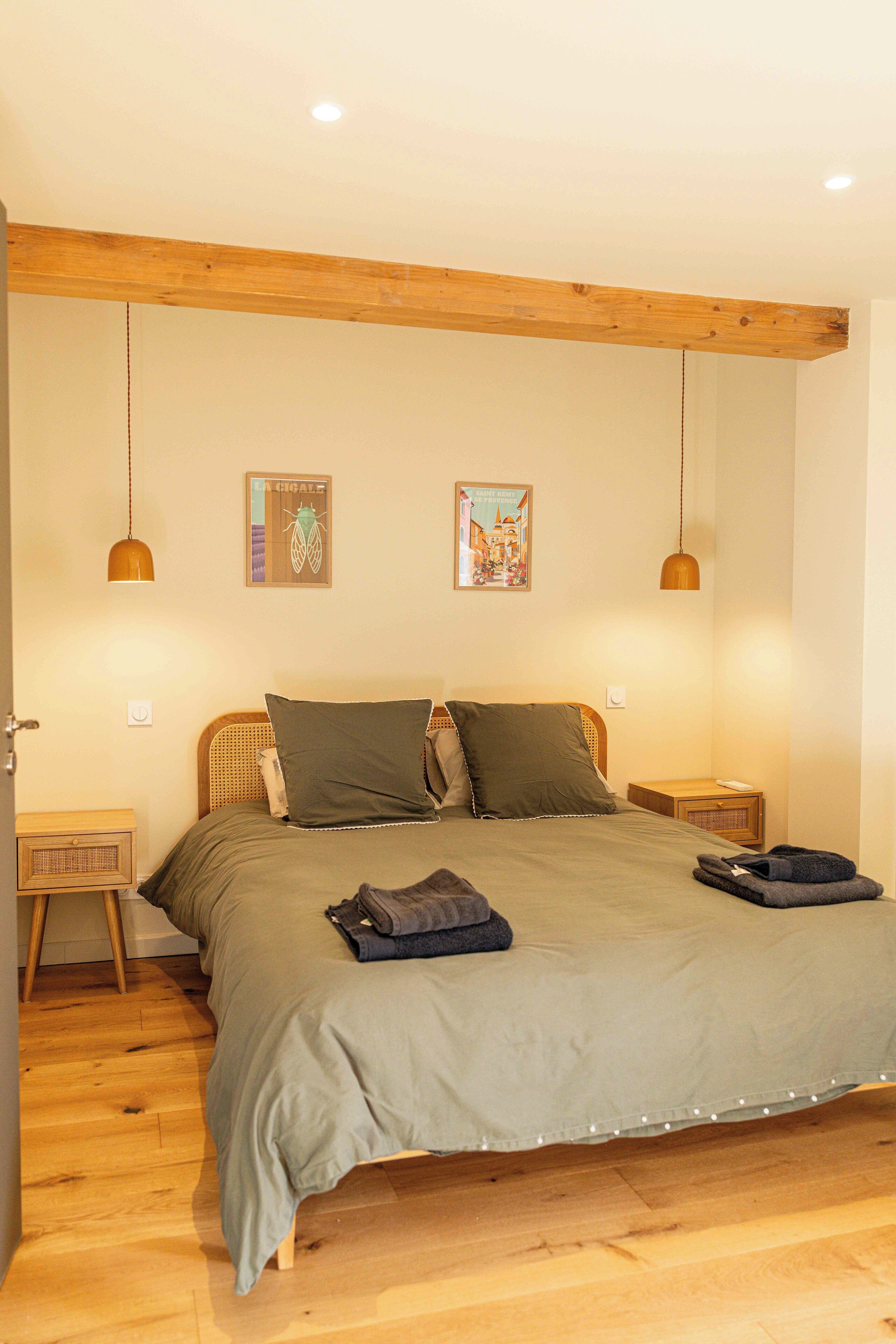 A bedroom with a wooden ceiling, two bedside tables, a bed with a green bedspread, two pillows, and two wall-mounted pendant lights.