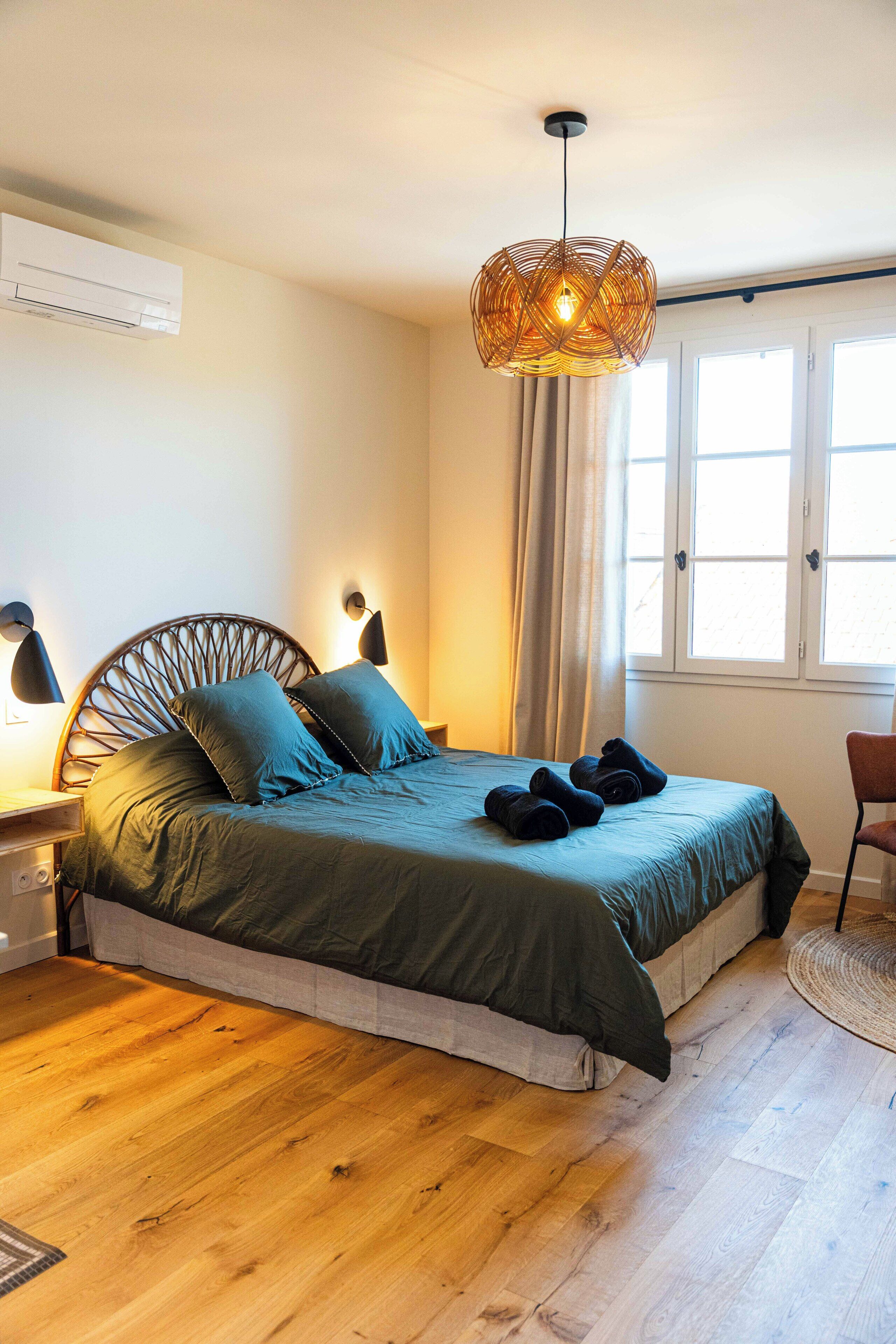 A bedroom with a bed, bedside lamps, a hanging light fixture, a window with curtains, and a wooden floor.