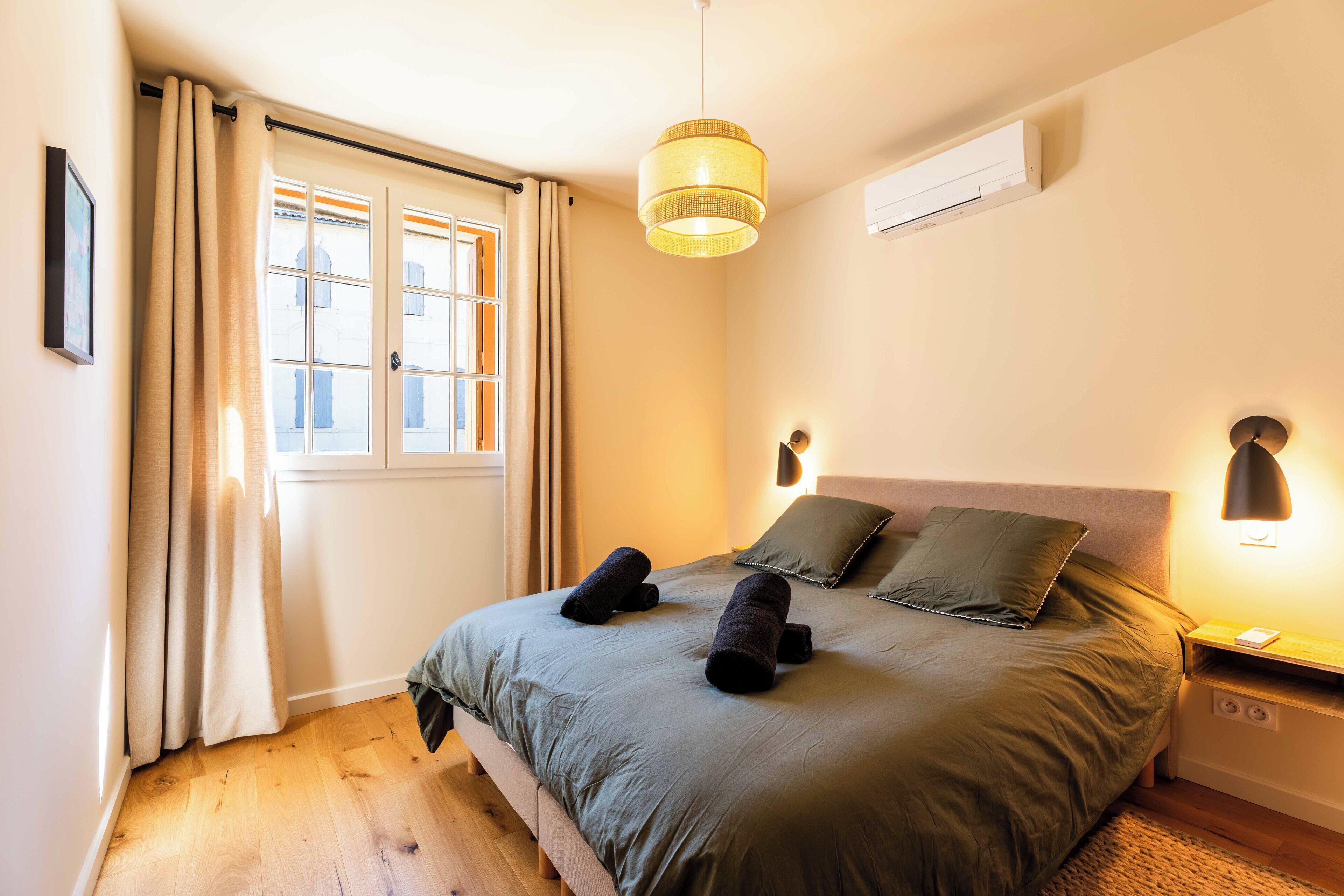 A bedroom with a bed, a window with curtains, a hanging light fixture, and a bedside table with a lamp.