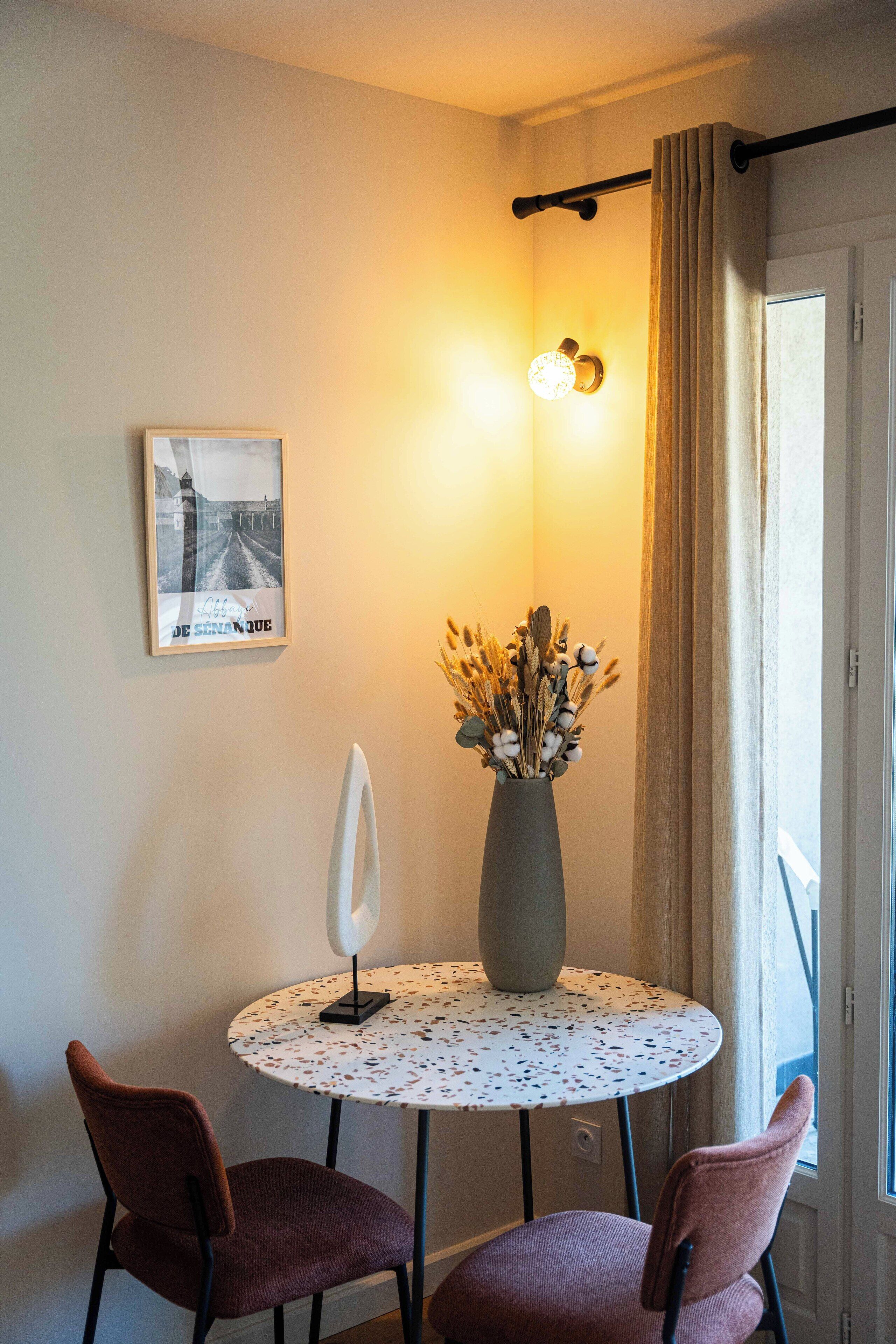 Un tavolino rotondo con un vaso di fiori e due sedie, un quadro appeso alla parete e una finestra con tende.