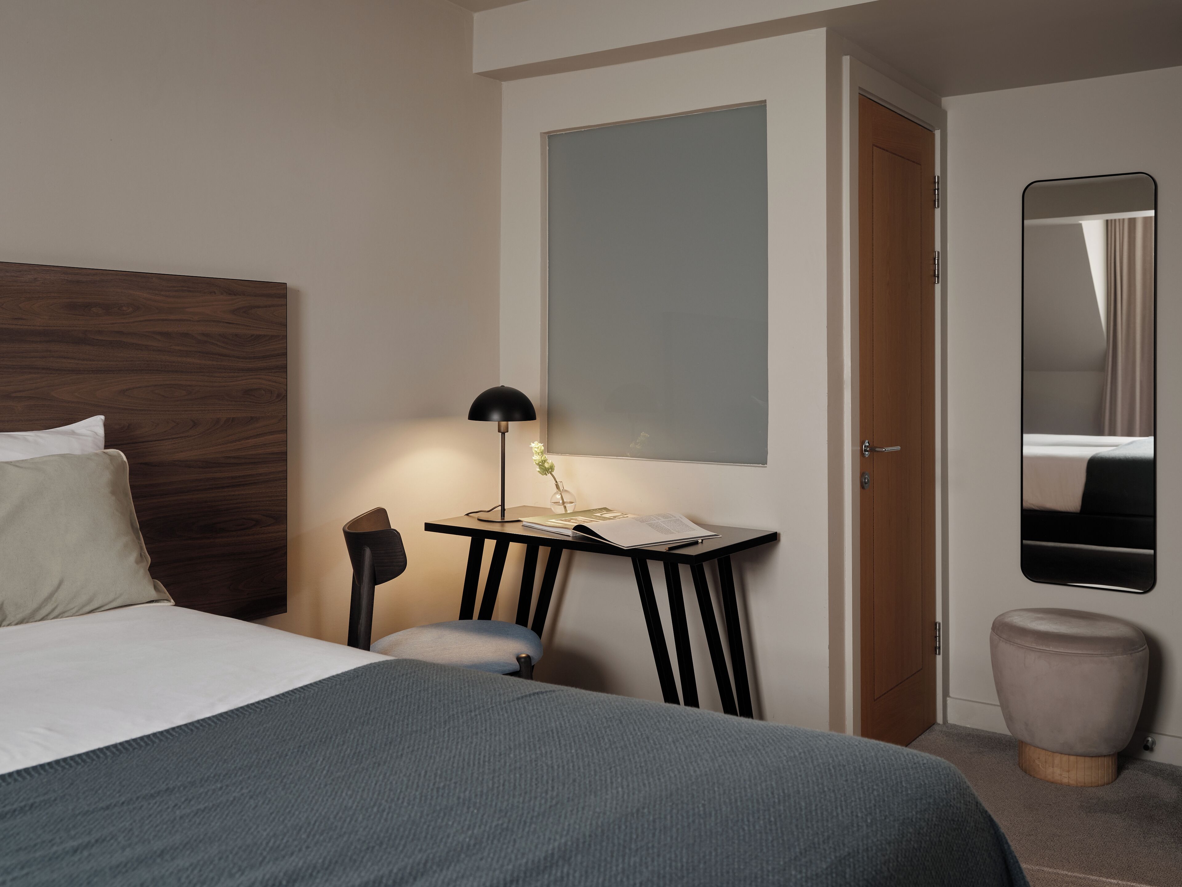 Et pænt møbleret hotelværelse med seng, skrivebord med lampe, spejl og en lille skammel.