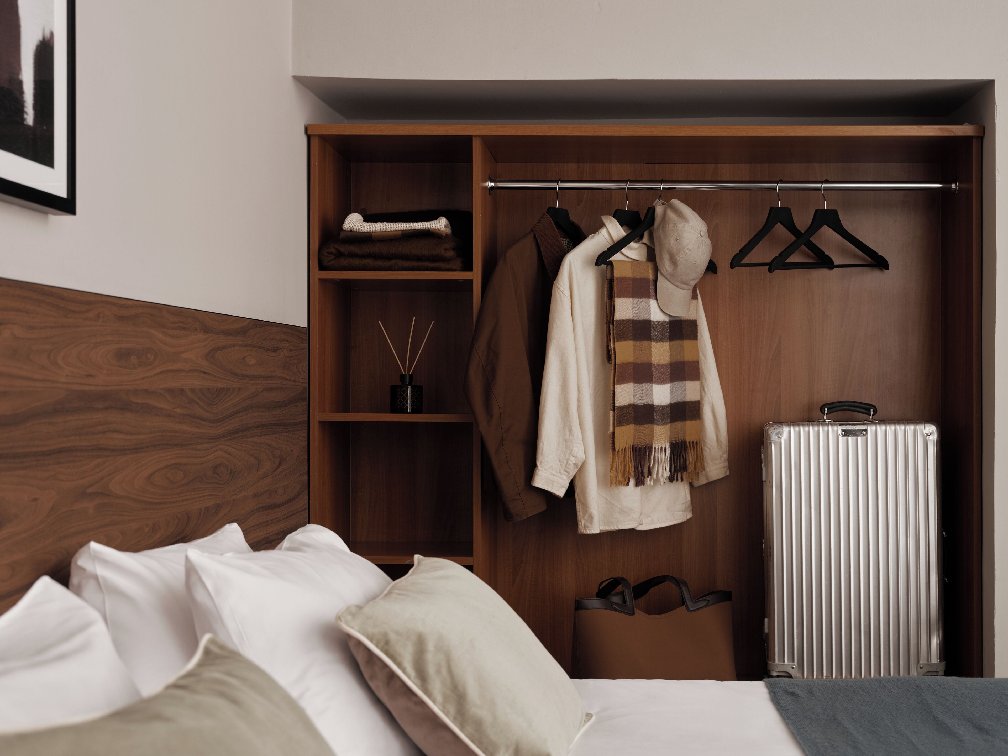 A neatly organized bedroom with a wooden headboard, a bed with white linens, a hanging coat rack with a scarf and hat, and a suitcase.